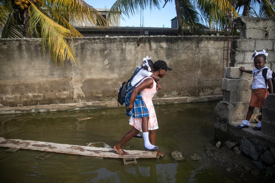 A daughter rides piggy back on her barefooted mother as they negotiate a flooded area caused by days of constant rain in Cap-Haitien, Haiti, Thursday, March 10, 2022.